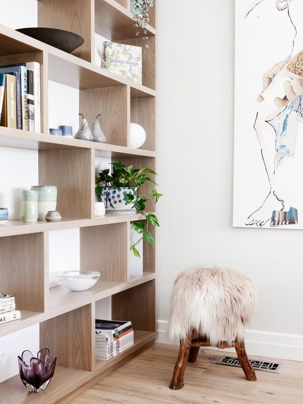 Bright, curated interior vignette featuring a full-height open timber shelving unit with asymmetrical cubbies displaying books, ceramics, glassware, plants, and decorative objects. A white wall with abstract artwork sits beside the shelving, while a shaggy cream-covered wooden stool rests on a light timber floor near a floor vent. The look is airy, warm, and contemporary with natural materials and soft neutral tones.