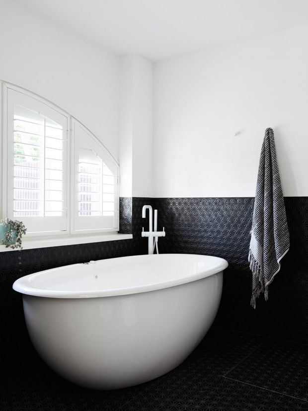 Bright contemporary bathroom featuring a freestanding white oval bathtub beneath a wall-mounted white tap, black patterned tiled lower walls and floor, white upper walls, and white shuttered windows. A striped towel hangs on the wall beside the bath.