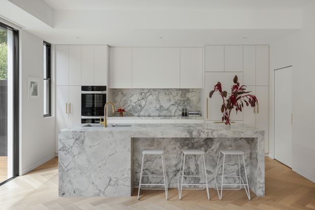 Bright contemporary kitchen with a large marble-look waterfall island, integrated white cabinetry, built-in black appliances, and a matching marble splashback. Three white wire-frame stools sit at the island, while pale timber herringbone flooring and a tall indoor plant add warmth.