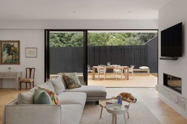 A bright contemporary living room with a large L-shaped upholstered sectional facing a wall-mounted television and built-in fireplace. Full-height black-framed sliding glass doors open to a timber deck with an outdoor dining setting, enclosed by a dark vertical privacy fence. The room includes pale timber flooring, a light neutral rug, nested coffee tables, restrained artwork, and a console, creating a refined indoor-outdoor connection.