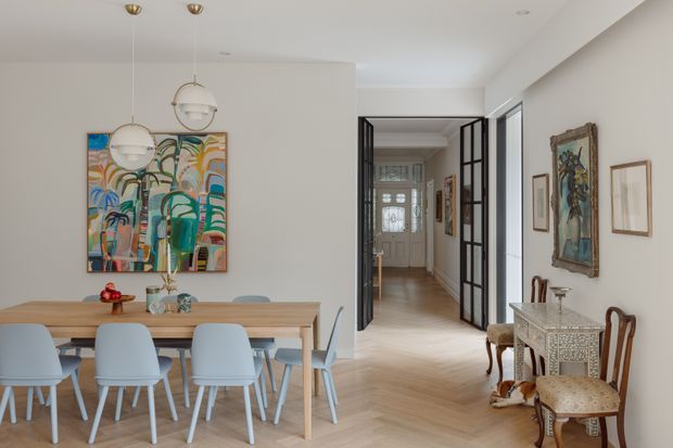 Bright open-plan dining and entry area with a light timber dining table, pale blue upholstered dining chairs, globe pendant lights, colourful abstract artwork, and light herringbone timber flooring. Black-framed glazed doors open toward a hallway and front entry, while an ornate console and antique-style chairs line the right wall.
