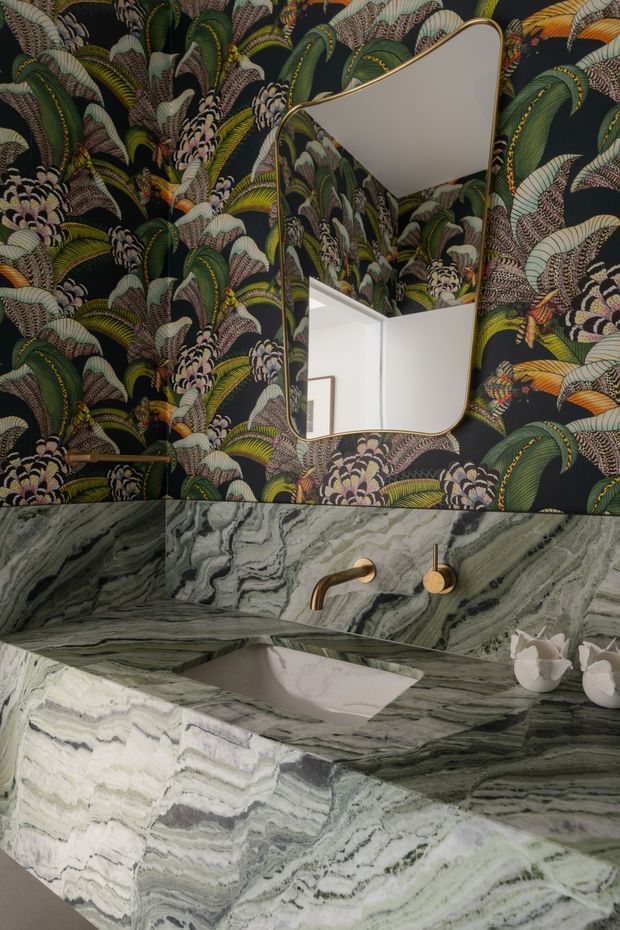 A close-up of a highly styled bathroom vanity. The walls are covered in dark, richly patterned botanical wallpaper, with an irregularly shaped mirror above a sculptural white basin. The vanity and backsplash are clad in dramatic green, grey, and white veined natural stone, paired with brushed brass wall-mounted tapware and a small white decorative vessel. The space feels bold, luxurious, and maximalist.