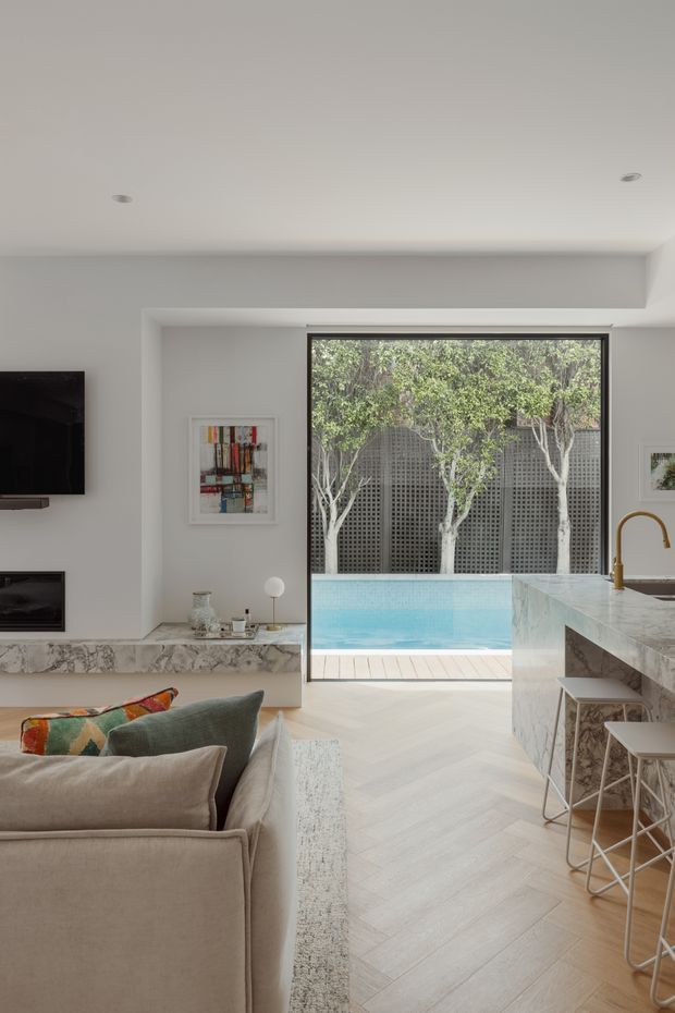 A bright open-plan living room and kitchen with a neutral contemporary interior. The room features a large sectional sofa, wall-mounted television and fireplace, marble-look hearth and kitchen island, pale herringbone timber flooring, brass kitchen tapware, and a large glazed opening overlooking a swimming pool and landscaped courtyard.