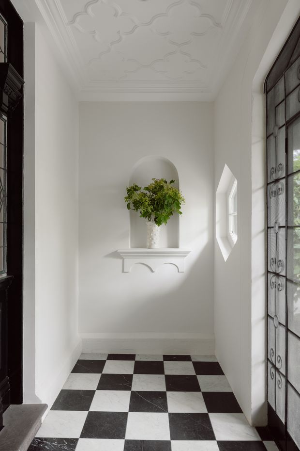 A narrow, elegant entry vestibule with white walls and ornate decorative ceiling moulding. The space features a black-and-white checkerboard stone or tile floor, a white arched wall niche with a small console shelf and floral arrangement, and tall dark-framed glazed doors or windows with decorative ironwork. Bright natural light enters from the right, creating a refined traditional atmosphere.