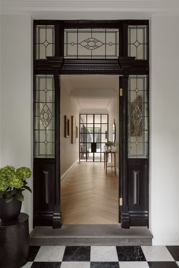 A formal entryway viewed through an ornate dark timber and stained-glass doorway. The hall beyond has a warm herringbone timber floor, white walls with decorative cornices and skirting, framed artwork, a console table, and a glazed internal door at the far end. The foreground entry floor is a dramatic black-and-white checkerboard tile pattern, with a potted plant to the side. The overall character is refined and heritage-inspired.