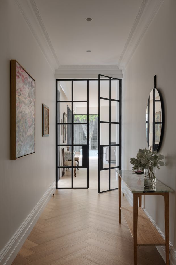 A refined residential hallway with pale herringbone timber flooring, white walls, ornate decorative ceiling cornices and recessed downlights. Black steel-framed glazed double doors open toward a bright courtyard or living area. A slim timber console with a light stone top, oval wall mirror and floral artwork add elegant finishing details.