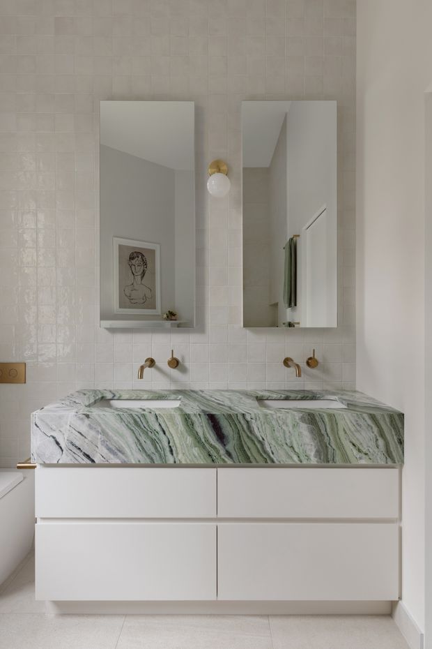 A refined bathroom vanity with a double basin, white flat-panel cabinetry, and a dramatic veined green-and-white stone benchtop. Two rectangular mirrors and brushed brass wall-mounted taps sit against a glossy pale tiled wall, with soft neutral finishes and minimal decorative styling.