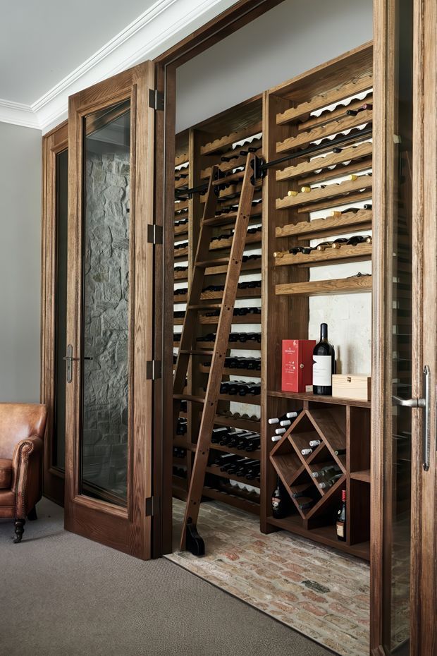A compact, richly detailed wine storage room viewed through open timber-framed glass doors. The room contains extensive built-in wooden wine racks filled with bottles, a rolling wooden ladder, a brick floor, and a small geometric bottle display. Dark-stained timber joinery and glass-panelled doors contrast with pale walls and carpeted flooring in the adjoining room.