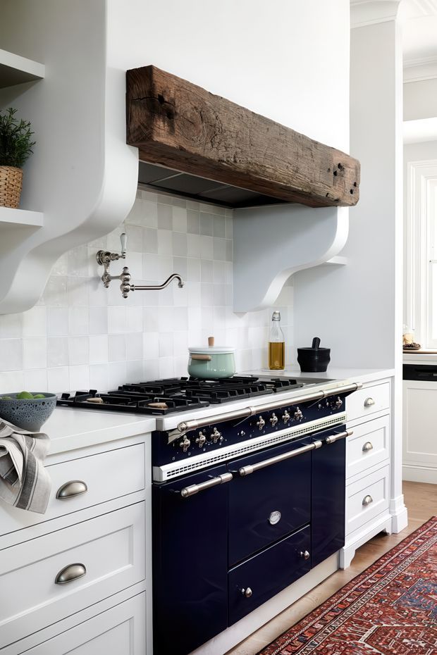 A refined traditional kitchen featuring a large navy-blue range cooker set within white Shaker-style cabinetry. The cooking zone has a glossy white square-tile splashback, a substantial reclaimed timber beam shelf over the range, sculptural white plaster hood surrounds, polished metal hardware, and warm timber flooring partially covered by a patterned runner rug. A glimpse of an adjoining bright room is visible through the opening on the right.