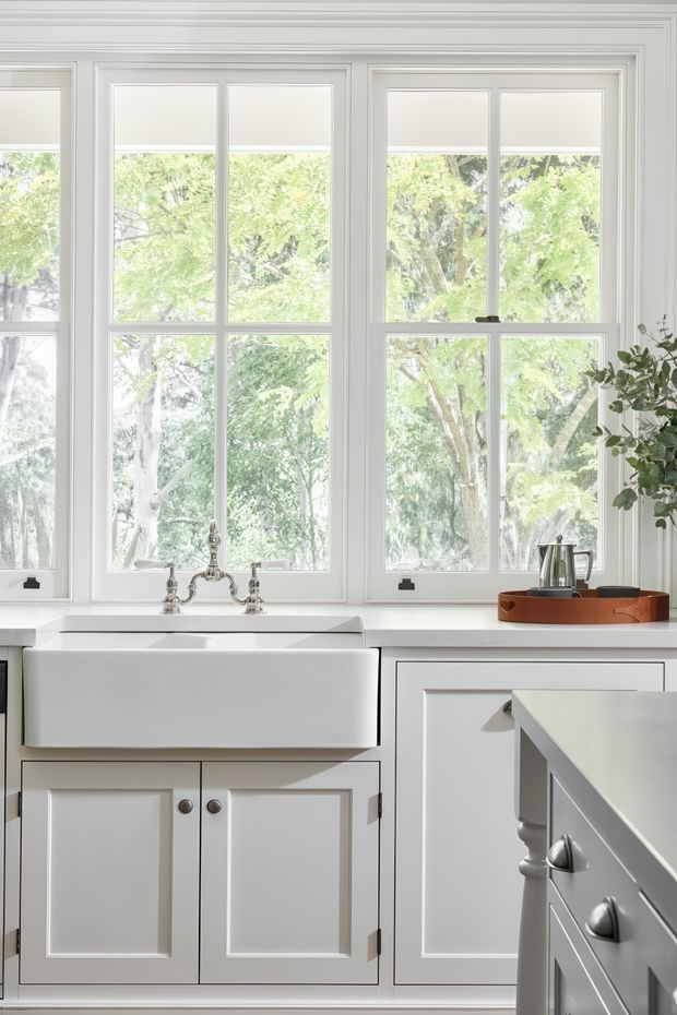 Bright, traditional-style kitchen with a deep white farmhouse sink beneath large multi-pane windows. White framed cabinetry with raised-panel doors, dark metal hardware, pale benchtops, and a partially visible island create a clean, classic composition. Lush leafy trees are visible outside the windows.