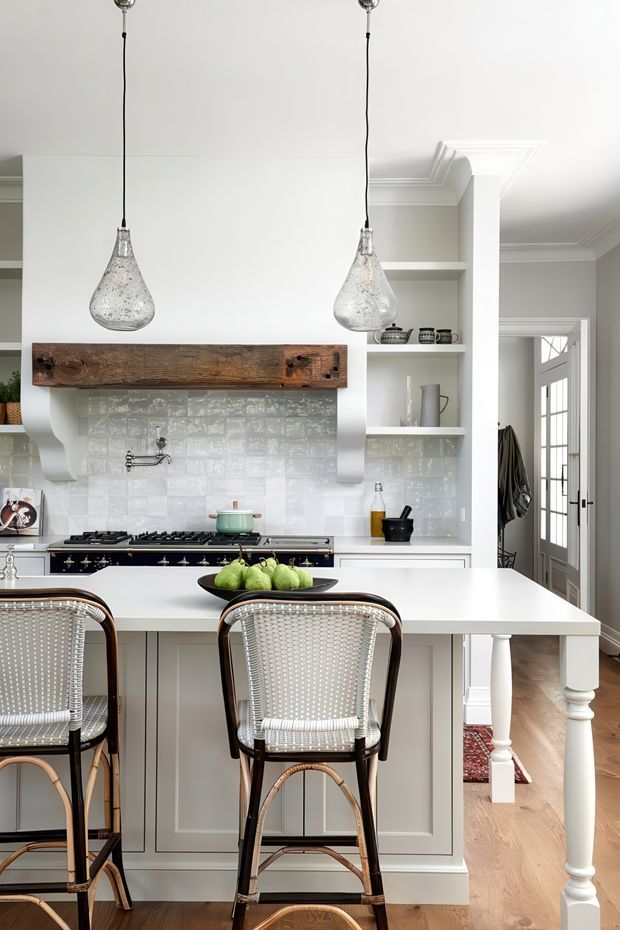 Bright, refined kitchen with a large white island and integrated seating, pale cabinetry, glossy textured white tile splashback, and a dark vintage-style range cooker. A reclaimed timber mantel shelf frames the cooking area, while glass pendant lights hang above the island. Open shelving displays ceramics, and a glazed door and timber flooring are visible to the right. The room combines classic architectural detailing with relaxed coastal styling.