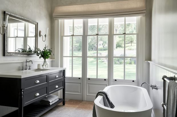 A bright, refined bathroom with a freestanding white bathtub positioned beside tall multi-pane French doors overlooking a garden. The room features a dark traditional-style vanity with a white countertop and basin, a large framed mirror, a white painted wall with decorative panel moulding, a radiator towel rail, and warm stone or tile flooring. Natural light and muted neutral finishes create an elegant, serene atmosphere.