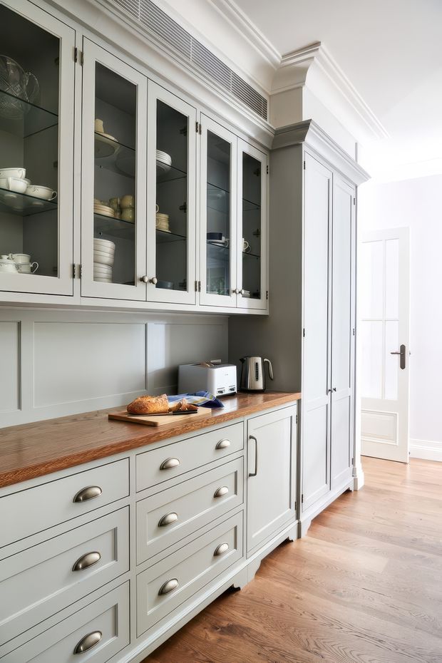 A bright, traditional-style kitchen butler's pantry or storage wall with pale grey-green shaker cabinetry, glazed upper cupboards displaying crockery, a warm timber benchtop, panelled splashback, and integrated tall storage. Chrome or brushed metal cup pulls and handles provide understated contrast. The room opens toward a bright adjoining area with a white paneled door, while wide timber floorboards continue throughout.