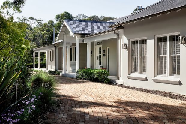Single-storey residential home exterior viewed from a landscaped brick-paved driveway or courtyard. The house has pale rendered walls, dark grey corrugated pitched roofs, white-framed multi-pane windows, decorative gable detailing, covered entry porches, and traditional veranda-like columns. Mature planting surrounds the approach.