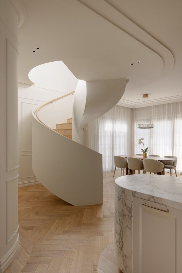 Elegant open-plan residential interior featuring a sculptural curved staircase with a smooth white balustrade, decorative wall panelling, a light herringbone timber floor, and a refined dining area with upholstered chairs and sheer curtains. In the foreground is a curved marble-topped kitchen island with pale cabinetry and brass hardware. The room has softly layered ceiling detailing and abundant natural light.