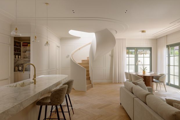 A bright, refined open-plan kitchen, dining and living space with an elegant curved staircase as the central feature. The room has extensive white wall panelling, a sculpted white ceiling, pale herringbone timber flooring, a large pale stone kitchen island with brass tapware, upholstered bar stools, a neutral sofa, and a compact round dining table beside tall glazed doors and windows. The overall design is softly contemporary with classic detailing and warm natural light.