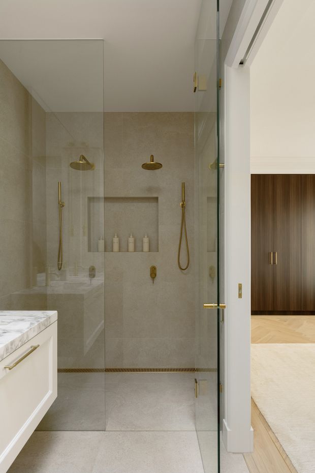 A refined contemporary bathroom centered on a frameless glass walk-in shower. The shower is lined with large-format warm light-grey stone-look tiles and includes dual brass showerheads, handheld sprays, recessed shelving with toiletries, and a linear floor drain. At left is a white built-in vanity with a pale veined stone benchtop and brass hardware. Through the open glass door, a warm timber-clad built-in cabinet and pale timber flooring are visible in the adjacent room.