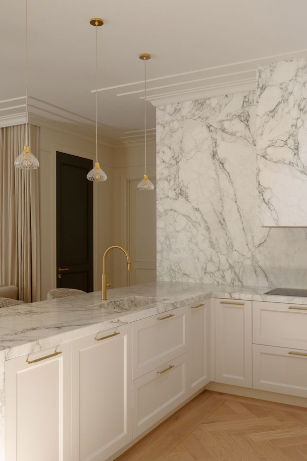 A refined, warm-toned kitchen with cream Shaker-style cabinetry, brass pulls and faucet, a large white veined marble backsplash and benchtop, and a light timber herringbone floor. The space has ornate ceiling mouldings, full-height curtains, dark timber internal doors, and three crystal pendant lights, creating an elegant classic interior.