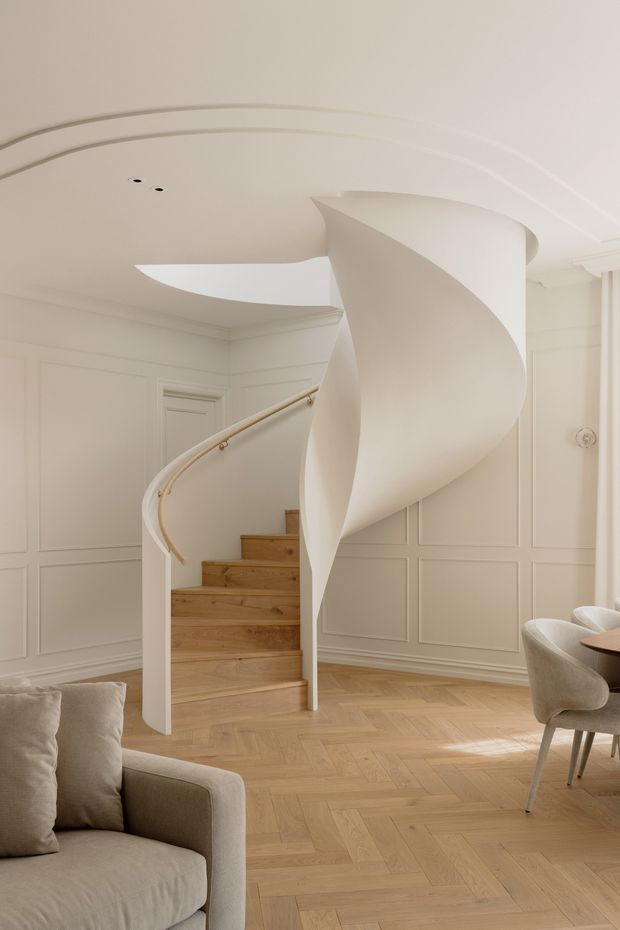 A refined residential interior featuring a sculptural white spiral staircase with warm timber treads and a slim curved handrail. The staircase rises through a dramatic curved opening in a white ceiling, set against elegant cream wall panelling and ornate ceiling mouldings. A pale herringbone timber floor, neutral upholstered sofa, and curved dining chairs create a calm, luxurious and cohesive interior.