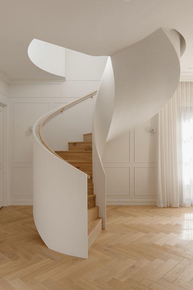 Interior view of a sculptural curved spiral staircase with a smooth white plaster balustrade and overhead sweeping form. The staircase has warm timber treads and a slim brass-toned handrail, set against white decorative wall panelling, sheer curtains, and a light herringbone timber floor. The composition is bright, refined, and minimalist with classical detailing.