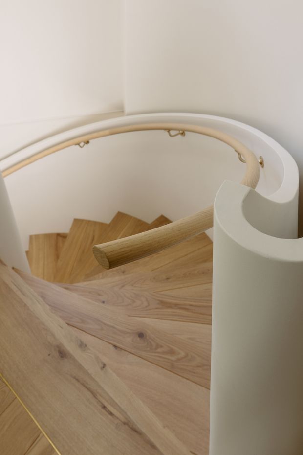 Close-up architectural photograph looking down a curved staircase with warm timber wedge-shaped treads, a rounded white stair enclosure and balustrade, and a pale timber handrail. The composition emphasizes the sculptural spiral geometry and natural wood grain in a softly lit, minimal interior.