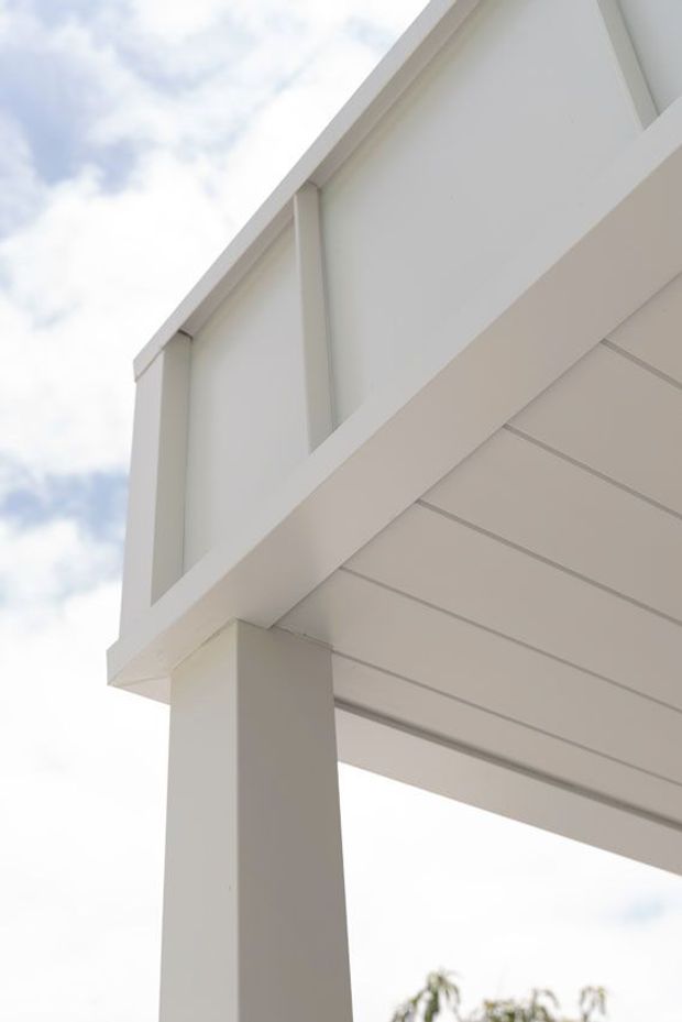 Close-up upward view of a clean white residential exterior, showing a gabled roof edge, broad fascia and eaves, horizontal soffit lining, and a substantial square support post. The image emphasizes crisp minimalist detailing and white painted surfaces against a partly cloudy sky.