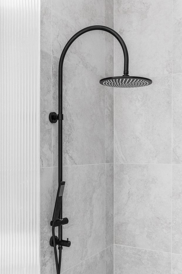 A close-up view of a contemporary shower enclosure featuring a matte black exposed shower system with a curved riser, large round rain showerhead, handheld shower, and controls. The walls are clad in large-format light grey stone-effect tiles, with a narrow white vertically ribbed feature panel at the left edge.