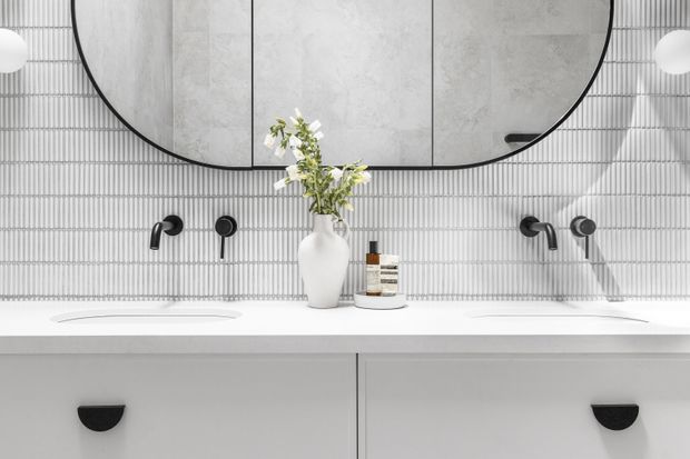 A refined contemporary bathroom vanity with two white basins, black wall-mounted tapware, pale grey cabinetry, and a vertically ribbed light tile splashback. A large rounded black-framed mirror spans the vanity, with a white vase and small toiletries arranged on the counter. The palette is predominantly white, light grey, and black with a clean, symmetrical composition.