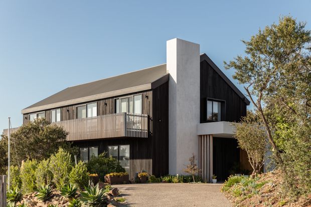 Exterior view of a contemporary two-storey gabled home with dark vertical timber cladding, a light rendered chimney or central wall element, extensive glazing, and a projecting upper-level balcony. The house is set within a landscaped garden with a paved driveway and established planting.