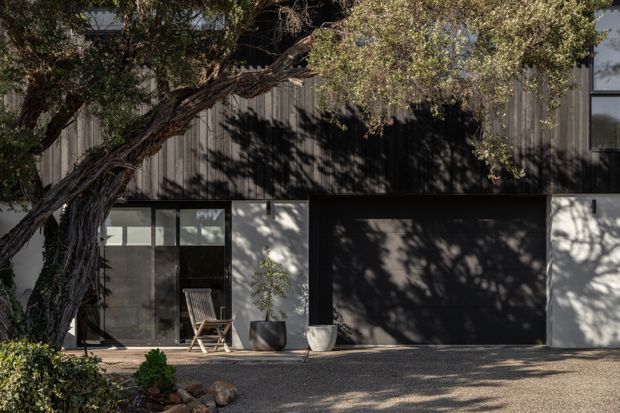 Exterior view of a contemporary multi-level residence, featuring dark vertical cladding on the upper facade, pale rendered lower walls, a large black garage door, and glazed entry doors. A mature tree casts shadows across the facade and gravel forecourt.