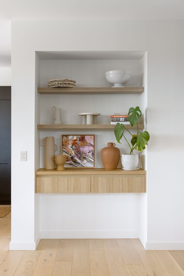 A built-in recessed display niche with three timber shelves and a matching floating cabinet, set against white vertical wall panelling. The shelves display neutral-toned ceramics, books, artwork, woven objects, and a potted monstera. Pale timber flooring and crisp white walls create a calm, light-filled contemporary interior.