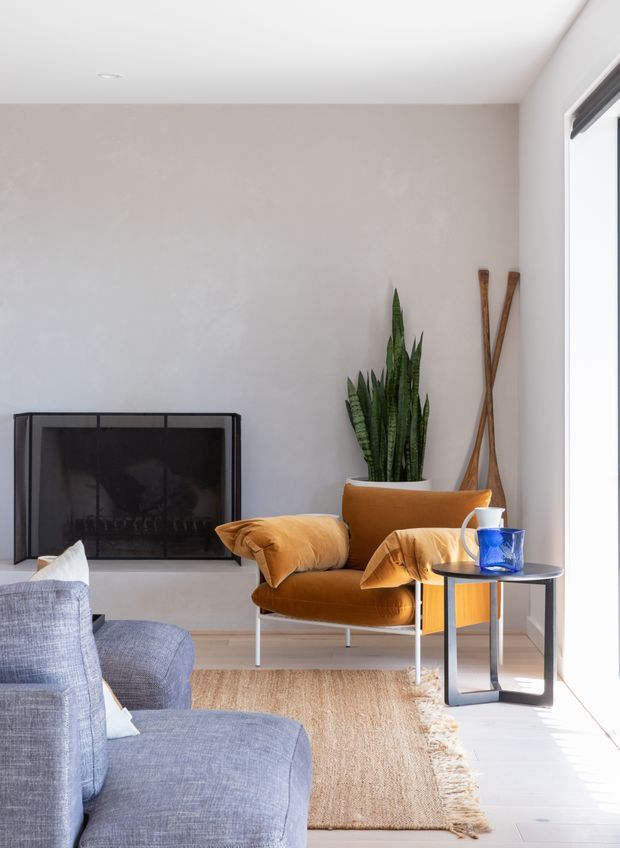 A bright, contemporary living room with a neutral plastered wall, white ceiling, large window, and a black-framed fireplace. The room features a caramel velvet lounge chair with matching cushions, a blue-grey sofa in the foreground, a dark metal side table, a tall snake plant, decorative wooden objects, and a natural woven rug. Sunlight creates strong illumination along the right side of the room.