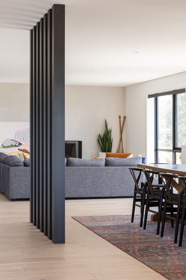 Bright open-plan interior showing a dining area and living room divided by a striking floor-to-ceiling black vertical-slat screen. The space has pale timber flooring, white walls and ceiling, a grey upholstered sofa, dark dining furniture, a large patterned rug, indoor plants, and generous black-framed glazing. The style is clean and contemporary with a strong architectural feature partition.