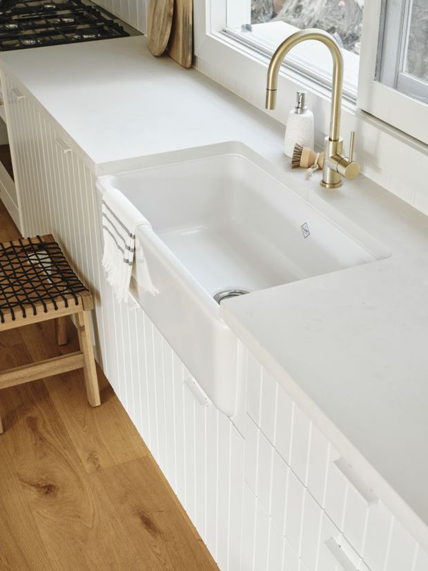 Bright close-up of a white kitchen featuring a deep farmhouse-style sink integrated into white cabinetry with vertical fluted detailing. The kitchen has a pale solid-surface benchtop, brushed brass gooseneck tapware, a black gas cooktop, timber flooring, and a timber stool with woven seat beside the cabinetry. Natural light enters through the window.
