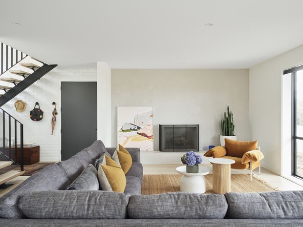 Bright contemporary living room with a large grey sectional sofa, warm timber flooring, sculptural coffee and side tables, mustard accent cushions and lounge chair, a black-framed fireplace, and a minimalist open staircase with black metal balustrade. White walls, tall glazing, and a tall indoor plant create an airy, refined interior.