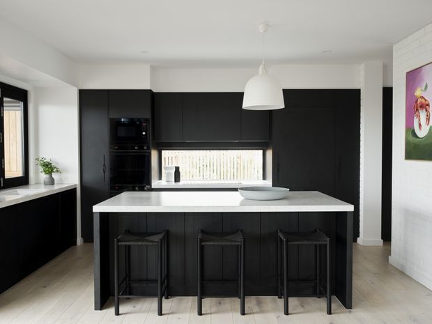 A contemporary kitchen with a large white stone island, three black bar stools, and extensive matte-black cabinetry. The room has pale timber flooring, white walls, a white pendant light, integrated appliances, and a horizontal window above the rear benchtop. A white-painted brick wall and colourful artwork are visible on the right.