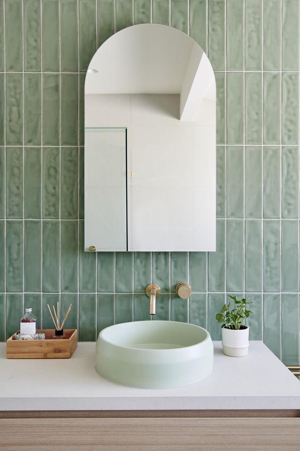 A refined contemporary bathroom vanity featuring vertically arranged sage-green glazed tiles, a pale stone benchtop, a rounded pale green vessel basin, brushed brass wall-mounted tapware, and a large arched mirrored cabinet. Timber-look vanity cabinetry, a small tray of toiletries, and a potted plant add warmth and texture.