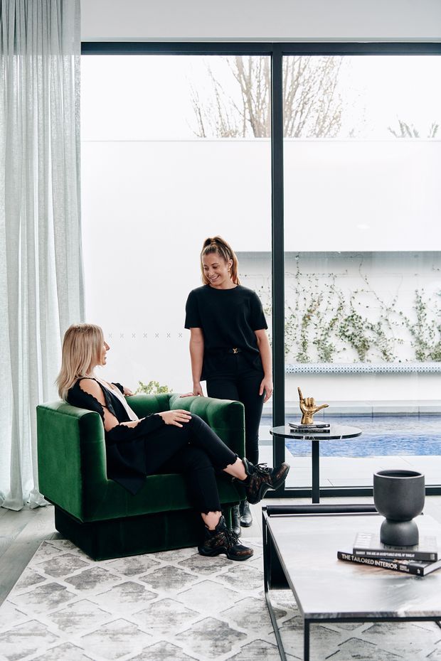 Contemporary living room with a deep green upholstered armchair, black-clad seating area, slim black coffee tables, patterned rug, sheer floor-to-ceiling curtains, and large dark-framed glazing overlooking a pool courtyard. The room has a refined monochromatic palette with green upholstery accents and abundant natural light.