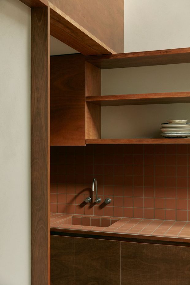 Close-up view of a compact kitchen or kitchenette with dark timber framing and cabinetry, open timber shelves, a terracotta-red tiled backsplash and tiled benchtop, and a small integrated sink with wall-mounted metal tapware. Stacked crockery sits on the shelving. The composition emphasises warm timber joinery, grid-pattern tiling, and built-in storage.