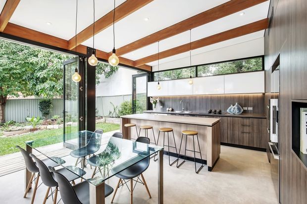 A bright open-plan kitchen and dining area with a large island, integrated cabinetry, glass dining table, and contemporary pendant lighting. Exposed timber ceiling beams contrast with white ceiling panels, while full-height glazing opens onto a landscaped garden. The kitchen features dark wood cabinetry, a pale island benchtop, and a narrow horizontal clerestory window.