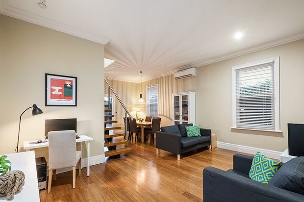 Bright, compact open-plan living and dining area with polished timber flooring, warm neutral walls, white trim, and a sculptural staircase leading to an upper level. The room includes a small desk and chair, dining table, dark sofas with colourful patterned cushions, a glass-front cabinet, plantation-style blinds, and recessed lighting. A large wall opening visually connects the living area with the dining and stair zone.