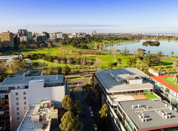 Aerial view over a dense urban district beside a large landscaped park and lake. Modern mid-rise commercial and residential buildings occupy the foreground, while tree-lined streets, lawns, waterways and high-rise development extend toward the horizon.