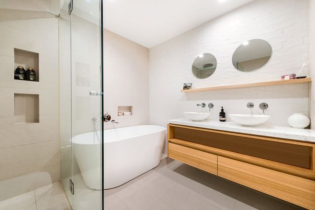 Bright contemporary bathroom with a freestanding white bathtub beside a frameless glass shower enclosure. A long floating timber vanity with two white vessel basins sits against a white painted brick-tile wall, topped by a timber shelf and paired round mirrors. The room uses light neutral finishes and warm timber joinery.