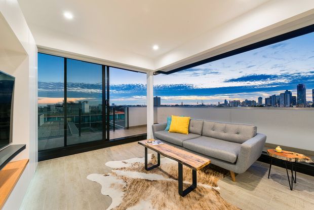 Bright contemporary apartment living room with floor-to-ceiling black-framed glazing and a balcony overlooking a waterfront city skyline. The room has a grey tufted sofa, timber coffee table with black metal legs, low media joinery, recessed ceiling lighting, and pale polished flooring. A yellow cushion provides a small accent of colour.