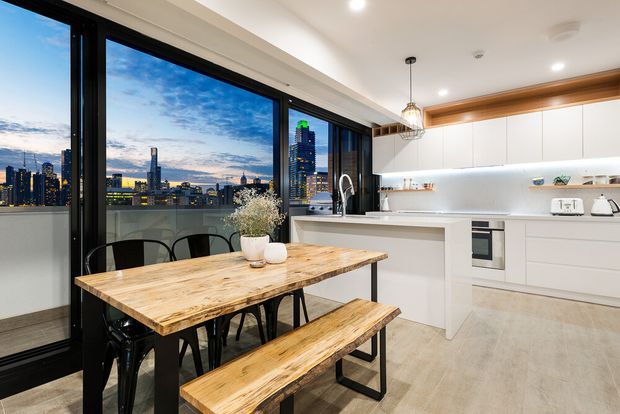 Bright contemporary kitchen and dining area with a large timber dining table and matching bench seating positioned beside expansive black-framed glazing. White flat-panel cabinetry, a pale stone-look island benchtop, integrated appliances, open shelving and under-cabinet lighting create a clean, modern fit-out. The glazing overlooks a dense illuminated city skyline at dusk, while a pendant light hangs above the dining area.