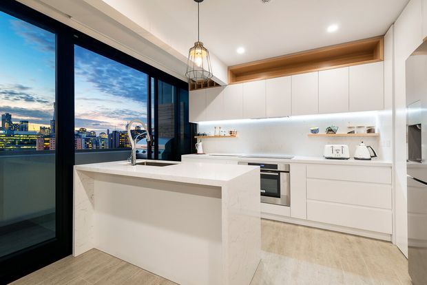 Bright contemporary kitchen in an apartment, featuring a white waterfall-edge island with integrated sink, extensive white flat-panel cabinetry, a pale stone benchtop and splashback, timber-look flooring, and a warm timber open shelf above the upper cabinets. Full-height black-framed glazing opens to a balcony and overlooks a dense city skyline. Pendant and recessed lighting provide layered illumination.