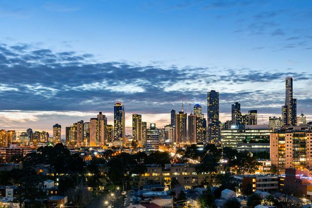 Twilight cityscape showing a dense modern urban skyline with numerous illuminated high-rise residential and commercial towers, viewed from an elevated suburban area. Dark tree canopies and low-rise buildings occupy the foreground beneath a blue, cloud-filled evening sky.