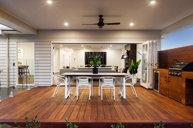 A bright, open-plan entertaining area with a timber deck, white dining table and chairs, and a connected living room visible beyond wide open glazing. The space features white horizontal wall cladding, warm timber flooring and cabinetry, recessed ceiling lights, a ceiling fan, indoor plants, and a built-in outdoor kitchen or barbecue along the right wall.