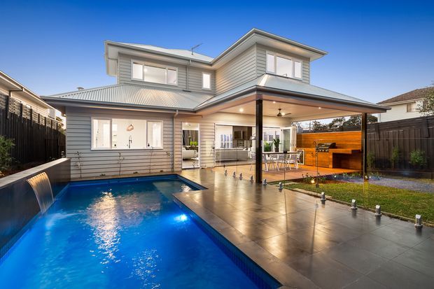 A two-storey suburban home with light horizontal weatherboard cladding and a pitched metal roof, viewed from the rear. The outdoor area features a large rectangular swimming pool, paved entertaining terrace, covered patio with dining furniture, built-in timber outdoor kitchen or barbecue area, lawn, and boundary fencing. The scene is photographed at dusk with exterior lighting reflecting in the pool.