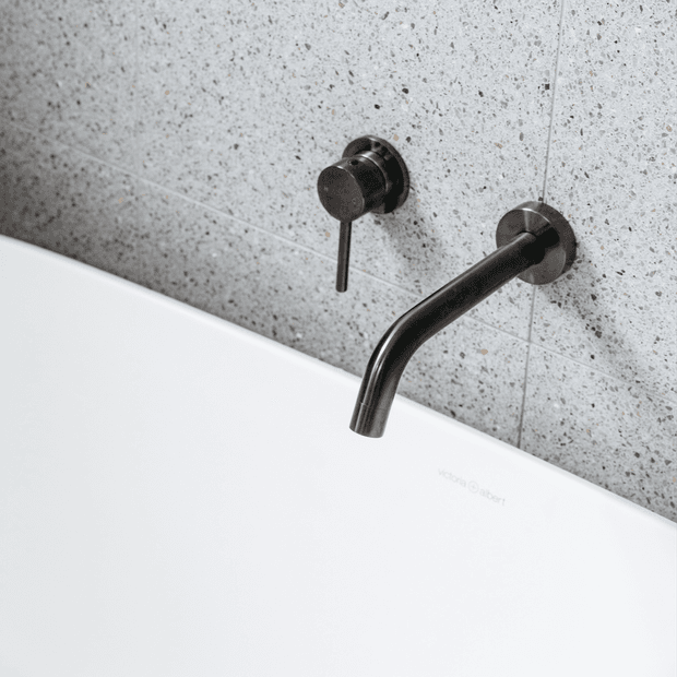 Close-up of a minimalist bathroom wall-mounted bath filler and control in a dark brushed metal finish, positioned above a white freestanding bathtub. The wall is clad in light grey speckled stone-look tiles with fine grout lines.